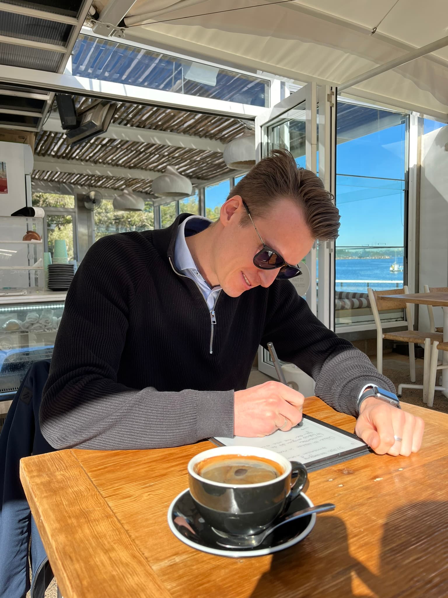 Xander writing notes at a waterfront cafe over coffee