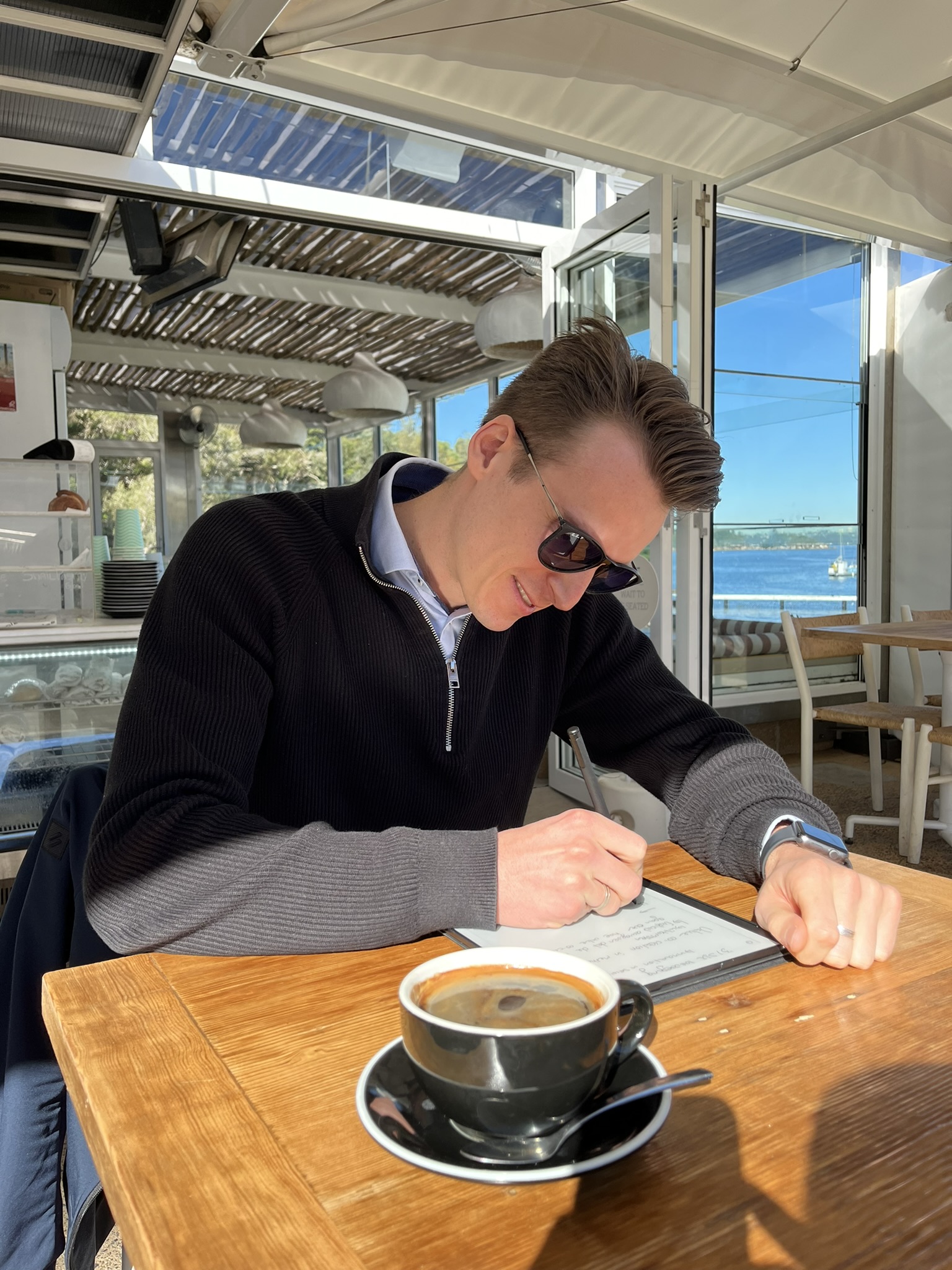 Xander writing notes at a waterfront cafe over coffee
